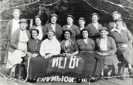 Photograph of fourteen middle-aged women posed in front of trees; they are wearing hats with a badge on the front and cardigans, also bearing a badge, and dresses or skirts; the women sitting on the front row have a cloth over their knees and the woman in the middle is holding a trophy cup; five bowling balls are lying on the ground at their feet; they have been identified as the founder-members of the Ladies' Bowling Club in Wingate