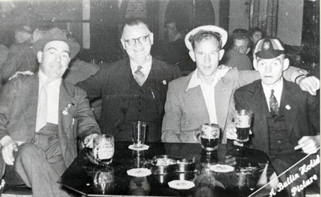 Photograph showing four men sitting at a small table on which there are three glasses and an ashtray; behind the men other groups can be seen indistinctly; the man at the left of the table is wearing a suit, tie and trilby hat; next to him is a man wearing a suit and waistcoat and glasses; next to him is a younger man wearing an open-necked shirt and a jacket; the man on the right is wearing a suit and tie and waistcoat and what appears to be a school cap; in the corner of the photograph are the words: A Butlin Holiday Picture; the photograph has been identified as Mr. Bell (of Wingate) at Butlins