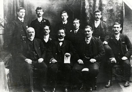Photograph of ten men wearing suits, stiff collars and ties, posed in a group in front of curtains; a man in the middle of the front row is holding an illegible piece of paper; they have been identified as Staff of Prudential Insurance