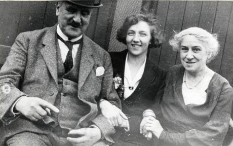 Photograph of a middle-aged man wearing a suit, wing collar, tie, waistcoat, and hat sitting in front of a wooden fence or shed; a young woman wearing a jacket and blouse has her arm through that of the man; a middle-aged woman wearing a frock and a necklace has her arm through that of the young woman; they have been identified as Dr. Willie Arthur and his family