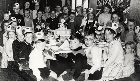 Photograph showing twenty four children, aged approximately between two and ten years, wearing party hats and sitting round a table, on which there are plates and a cake with a crown on top; six women are standing at the back of the children; on the wall behind the children are a Union Jack and a portrait of Queen Elizabeth II; they have been identified as taking part in Coronation Celebrations at Wingate