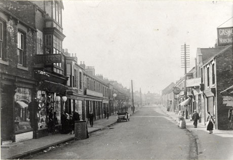 Photograph showing a road running away from the camera; on either side are shops; on the left are W. Harland, Fruiterer, and King's, possibly a draper's shop; the other shops on the left cannot be identified; on the right a branch of Walter Willson can be identified, but no other shop can be identified; a motor car is parked in the road facing away from the camera; two barrels are standing in the gutter outside a shop on the right; indistinct figures are walking and standing on the pavements; the street has been identified as Front Street, Wingate