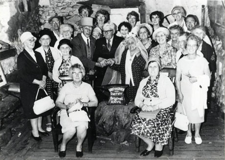 Photograph of twenty three elderly people standing round a stone on which there is an anvil on which the following is written: Gretna Green Smithy Original Anvil; behind the group are rough stone walls; an elderly man is dressed as a bridegroom and an elderly woman is dressed as a bride and another elderly man is holding their hands together over the anvil; the group has ben identified as the Over 60s Club in Wingate on outing to Gretna Green