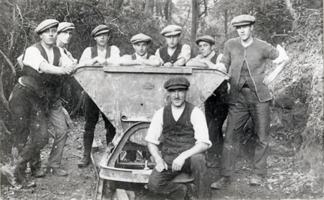 Photograph showing eight men standing round a machine with trees and bushes behind and on either side of them; the machine is unidentifiable; the men are wearing work clothes of trousers, waistcoats and caps; the photograph has been identified as Preparing The Running Track, The Planting, Wingate