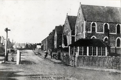 Postcard photograph entitled Church Street, Wingate, showing a level crossing in the foreground with a street beyond, running away from the camera; on the left are indistinctly seen fronts of houses; on the right are the signal box of the railway, the front and side of what appears to be two chapels, and, beyond them, the front of a detached house and terraced houses; an indistinct woman and group of children can be seen on the pavement on the right