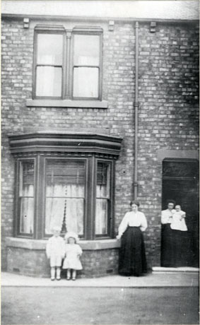 Photograph of the front of a terraced house showing a bay window, doorway, and window on the upper floor; in front of the bay window a small boy, aged approximately six years, is standing with a small girl, aged approximately four years; a woman, dressed in a light-coloured blouse and a long dark skirt, is standing near the window; a woman, similarly dressed, is standing in the doorway holding an infant; they have ben identified as Mrs. Bell and Child