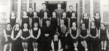 Photograph showing eighteen girls in gym slips, aged approximately fourteen years, grouped with two men wearing academic gowns sitting on the front row; three men and two boys, aged approximately fourteen years, are standing at the back of the group; the entrance and part of the walls and windows of the building behind them can be seen; they have been identified as Pupils in front of A. J. Dawson Secondary School, Wingate