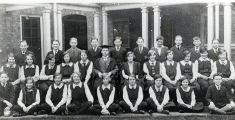 Photograph of seventeen girls, aged approximately fourteen years, posed with twelve boys of the same age in front of what appears to be the covered walkway of a building; the girls are wearing gym slips and the boys blazers; a man and a woman wearing academic gowns are sitting in the middle of the middle row; the group has been identified as A. J. Dawson Grammar School, Wingate