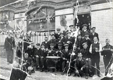 Photograph of thirty one men sitting and standing on the pavement outside an unidentified public house; the name of the licensee can be seen above the doorway, as follows: Tom Stewart, Licensed To Sell Beers, Wines, Spirits, and Tobacco; the exterior of the public house is tiled and has streamers and crowns decorating it; the words God Save The King can be seen in a window; the men are grouped round a wrought iron table and are holding glasses and mugs; a man on the right is holding an accordion; the photograph has been identified as Queen Mary and George V's Coronation, Wingate