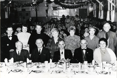 Photograph showing a table laid with cutlery, crockery and food, running across the front of the photograph, with four men and one woman sitting at the table facing the camera; four women, two men and a clergyman are standing behind them facing the camera; behind the people at the front of the picture, tables running away from the camera the length of the room with people sitting at them can be seen; two of the men are in dinner jackets; the other men are in suits; the women are in dresses and hats; they have ben identified as Over 60's Christmas Party, Wingate
