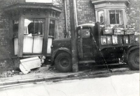 Photograph showing a bay window of a terraced house with the wall beneath the window in pieces and the window pushed to one side by a lorry, which can be seen on the pavement in front of the house with its bonnet in the window; there are boxes in the back of the lorry; the road in front of the house appears to be on a slope; the picture has been described as Ex-Army Truck Crashed into House (in Wingate) While Delivering Fish to North East