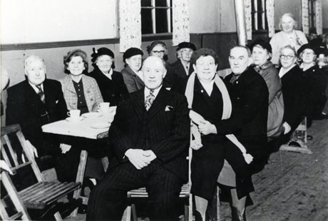 Photograph showing three elderly men and ten elderly women sitting either side of a long table; all the women are wearing overcoats and the men are wearing suits; one elderly woman is standing at the end of the table and she has been identified as Mrs. Champley; other members of the group have been identified as Mrs. Bell, Mrs. Ede; Mr. Purver; Mr. and Mrs. Cunliffe