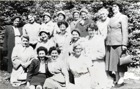 Photograph of sixteen middle-aged women dressed in suits, overcoats, dresses and hats, posed in front of trees; they have been identified as members of the Mothers' Union in Wingate on a trip