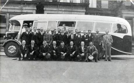 Photograph of twenty eight men posed in front of the side of a motor bus, which is parked in front of a building which appears to be a public house; a man in uniform, presumably the driver of the bus, and another man are standing in the entrance of the bus; the heads of two other men can be seen in the a window of the bus; the group has been identified as Buffs ROAB, Wingate