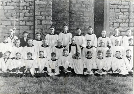 Photograph of fourteen men and fourteen boys, wearing surplices, posed against the wall of a church; a man carrying a processional cross, and a woman, dressed in a jacket and hat, are also on the back row of the group; a clergyman, dressed in surplice and preaching bands. is sitting in the middle of the middle row; the group has been identified as Church Choir, Wingate