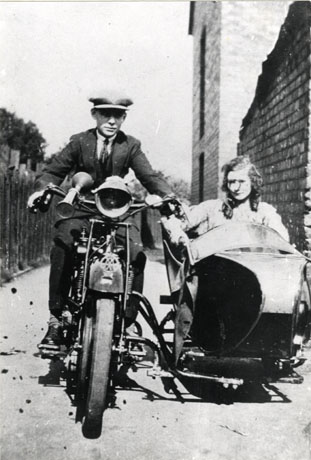 Photograph of a man, wearing a suit and cap, riding a motor bicycle, with solid tyres and bearing a badge of the Automobile Association, towards the camera; a side car is attached to the motor bicycle and woman can be seen sitting in it; she appears to be wearing a dress; a fence can be seen on the left of the picture and a wall on the right; they have been identified as Mr. and Mrs. Ord, Wingate