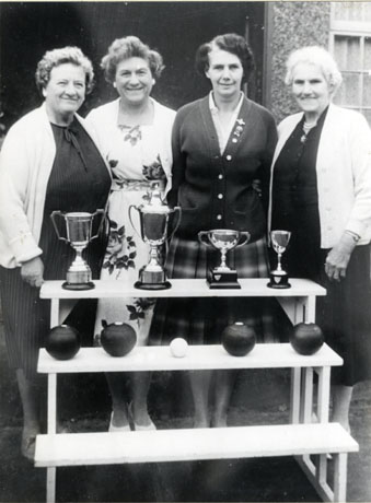Photograph of four middle-aged women standing behind a stand on which four trophy cups, four bowling balls and a jack are standing; the women are dressed in dresses, skirts and cardigans; the have been identified as Mrs. Bentham, Mrs. Rigby, Mrs. Bell, and Mrs. Brown, of Wingate