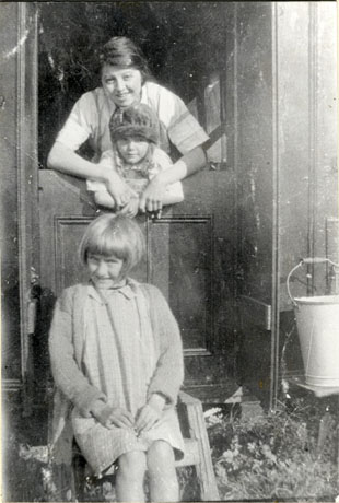 Photograph showing a woman leaning over a half door with her arms round a small girl aged approximately seven years; another girl aged approximately ten years is sitting in front of the door on a stool; a bucket is standing near the doorway at the right side of the doorway; they have been identified as Family at Bungalow at Tonks' Yard, Wingate