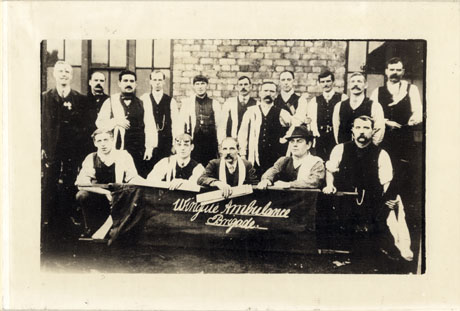 PHotograph of eighteen men posed in front of a brick building with a stretcher in front of them on which the words Wingate Ambulance Brigade has been written; most of the men are wearing trousers and waist coats but have removed their jackets; most of them have a cloth over their shoulder