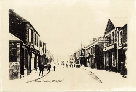 Postcard photograph, entitled Front Street, Wingate, showing a road running away from the camera with shops on either side; no shops can be identified on the left of the picture; only the Cash Clothing Co. The Workingman's Friends and Walter Willsons can be identified from their signs only on the right; unidentifiable people can be seen on the pavements