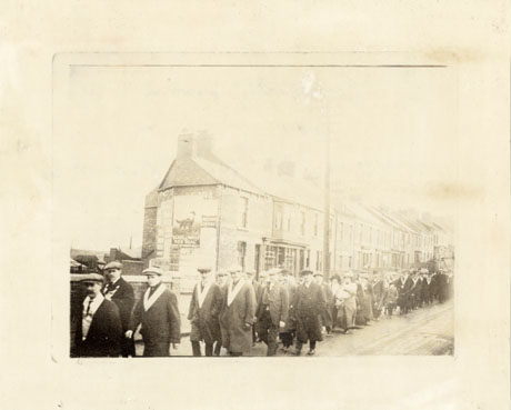 Photograph of a procession of men, women and children walking along an unidentified street past an unidentified shop and terraced houses; the men can be seen wearing insignia round their necks; the photograph has ben identified as Rechabites Procession to mark the opening of a social club in Wingate