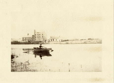 Photograph showing an indistinct individual in a rowing boat on a stretch of water in the foreground of the picture; across the water indistinctly, in the background, a tall building of four storeys with an attached building of two storeys can be seen; the photograph has been identified as Old Mill near Parr's Pond, Wingate