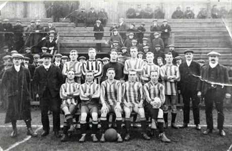 Photograph of twelve men in football strip posed with two other men in suits on either side of them; behind the group are the terraces of a football ground with men in suits and caps sitting on them; the photograph has been identified as Football Club, Wingate