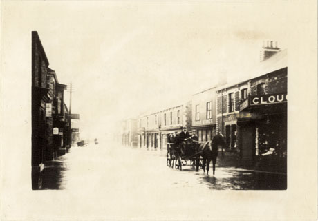 Photograph showing a road running away from the camera with shops on either side; the photograph is too light for the end of the road in the distance to be seen; on the right of the photograph can be seen a sign reading, Cycle Stores, and a sign reading, possibly, Clough; a horse and cart with, approximately, six indistinct passengers is approaching the camera and it can be seen that the surface of the road is covered in water to a depth that appears to cover the horse's hooves; the photograph is identified as The Flood, Wingate
