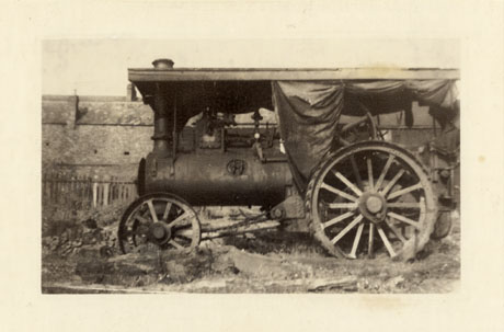 Photograph showing the side of a steam engine identified as Ted Dunlop's Threshing Machine; it is standing in what appears to be a dilapidated state in front of a fence with houses in the distance; it has two small wheels at the front and large wheels at the back; the cylinder and chimney for the steam can also be seen