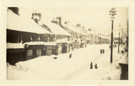 Photograph showing the side of a road with shop fronts nearest the camera and terraced houses further away; the road is covered so deep in snow that it is half way up the windows of the shops; the roofs of the buildings are covered in snow; indistinct figures can be seen in the road; the road has not been identified but it has been described as being in Wingate
