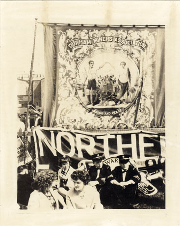 Photograph of the banner of Wingate Lodge of The Durham Miners' Association, showing a pair of allegorical figures representing Labour and Peace, the motto written on the banner; in front of the banner are two men in uniform sitting on the ground with brass musical instruments behind them; the head and shoulders of two women can be seen in front of them; the banner has been described as being at the Durham Miners' Gala