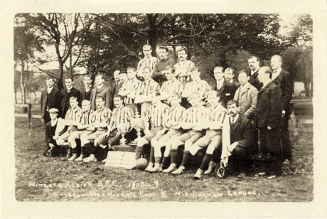 Photograph of fifteen men in football strip posed in front of trees at an angle to the camera; they are accompanied by thirteen men in suits; an inscription on the photograph identifies them as Wingate Albion Athletic Football Club 1906-7 Winners of Aged Miners' Cup Mid Durham League