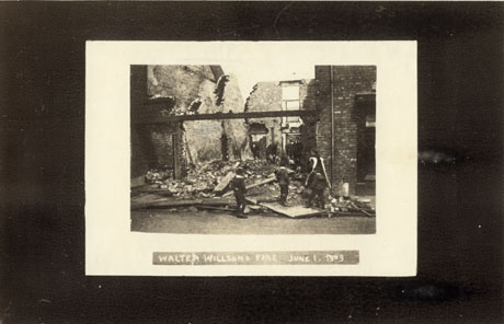 Photograph showing part of the front of a house with piles of bricks where the building next to it should have been; on the other side of the bricks the side of another building can be seen; the back wall of the damaged building can also be seen; three indistinct boys are standing in front of the ruin; a caption on the photograph describes the damaged building as Walter Willson's Fire, June 1, 1911; the shop has been identified as being in Wingate