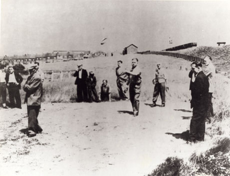 Photograph showing twelve men, wearing suits and caps, standing in a field with houses in the distance; all the men are looking towards something out of shot to the left; a man in the middle of the photograph has just thrown a quoit, which can be seen in the air in front of him; the photograph has been described as Bob Pounder Throwing Quoit; George Bryson Referee
