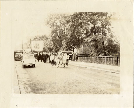 Photograph showing a procession walking towards the camera led by young girls followed by men carrying a miners' banner; the side of a house and a fence can be seen on the right of the picture; a motor car and a van can be seen on the road near the procession; neither the people in the procession nor the banners can be seen in any distinctness; the photograph has been described as Marching To Durham Miners' Gala, Wingate