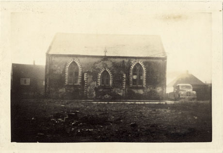 Photograph showing the side of a chapel with three pointed windows with coloured bricks outlining them; a car can be seen at the side of the chapel and two houses behind; in the foreground is open ground; the photograph is described as Chapel- Opened 1885, Wingate