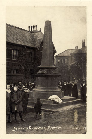 Photograph showing an obelisk surrounded by people and with buildings in the background; two young girls aged approximately twelve and thirteen are in the foreground; the obelisk has three steps surrounding it; the photograph bears a caption as follows: Wingate Disaster Memorial Dec. 21 1907