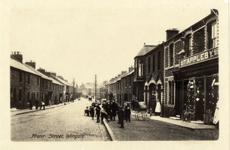 Postcard photograph entitled Front Street, Wingate, showing a road running away from the camera with terraced houses on the left and terraced houses at the far end on the right; a shop with the name, T. Appleby, above its window is on the right of the picture nearest the camera; its windows cannot be seen; indistinct figures are on the pavements and in the road; a horse-drawn vehicle is in the distance; the photograph has been identified as Front Street, Wingate