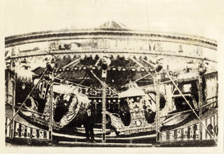 Photograph showing, indistinctly, the side of a fairground waltzer roundabout; two of the waltzer cars can be seen inside the structure and a man can be seen standing at the entrance to it; it has been described as Murphy's Showground, Wingate