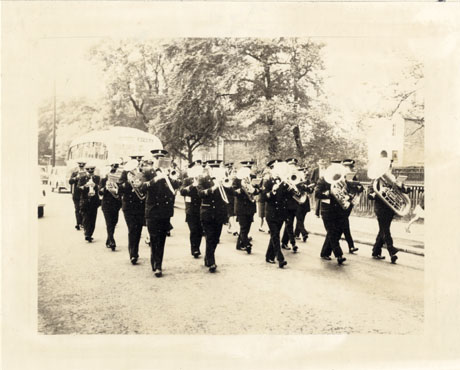 Photograph showing men in uniform walking along a road playing musical instruments; they are walking down the same road as the procession in wing0075; the top of a single-decker bus can be seen indistinctly behind the men; the photograph has been described as Yorkshire Band Playing and Marching, Wingate