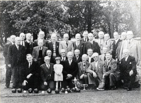 Photograph of a group of twenty nine elderly men standing and kneeling in front of trees; they are wearing suits and ties; a small girl aged approximately three years is standing at the front of the group; in front of the group are ten bowling balls and three jacks; the photograph has been described as Bowls League, Wingate