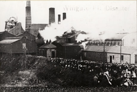 Photograph showing the buildings, chimneys and winding gear of a colliery with a crowd of people at the right of the picture; the photograph has the words Wingate Pit Disaster printed on it, so it is to be assumed that the crowd of people are waiting for news of the victims of the disaster; smoke appears to be coming from part of the pit