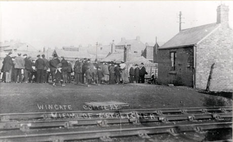 Photograph showing in the foreground railway lines running across the picture; beyond the railway lines are a crowd of people with their backs to the camera looking at something which cannot be seen; behind them are low buildings which may be houses; the photograph has the caption: Wingate Colliery View of Stretcher