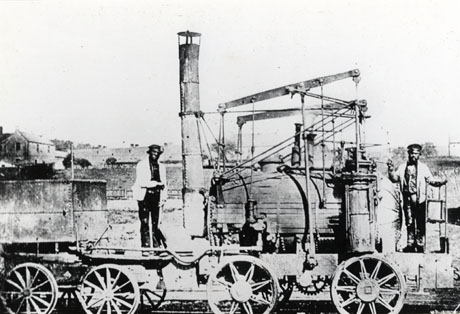 Photograph showing two men standing on a steam locomotive identified as Puffing Billy; the locomotive is seen from the side and is coupled to a truck; behind the locomotive are indistinct buildings; the men are also indistinct; the locomotive has been identified as being in Wingate