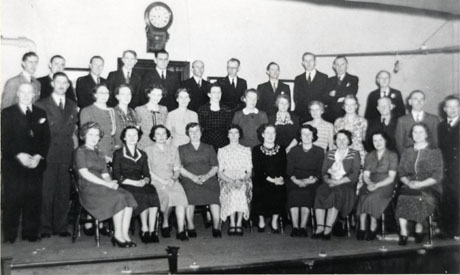 Photograph showing sixteen men in suits and ties, posed on a stage with nineteen women wearing dresses; behind them is a clock on the wall and a doorway; they have been unidentified, but is believed that they are in Wingate