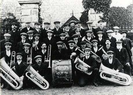 Photograph showing thirty five men wearing suits and caps and carrying brass musical instruments, posed outside the gateposts of a large house, part of the wall of which can be seen; they have been identified only as Brass Band, Wingate