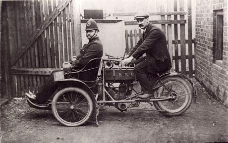 Photograph showing a man in the uniform of a police constable sitting in a seat at the front of a motor cycle with a man in a suit and cap sitting on the motor cycle at the back with his hands on the handle bars, which are behind the policeman; the frame of the motor cycle has the initials M. S. U. on it; the motor cycle and men have a fence and wooden gate behind them and a wall on the right; they appear to be in a back yard; thay have been identified as Ginger P. C. Neale and John Henry Stephenson in Wingate; the motor cycle has the registration number BB 218