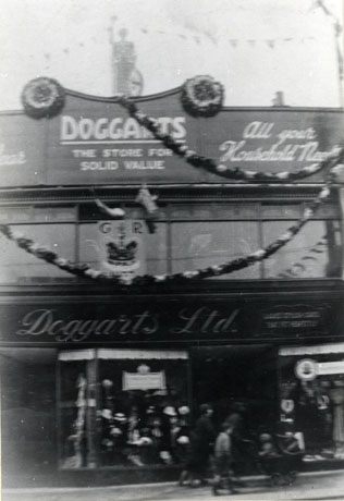 Photograph of the facade of Doggarts The Store For Solid Value All Your Household Needs decorated with steamers and with a crown and the letters G R( George Rex); a figure of Britannia can be seen on the roof of the shop; the windows and the people passing by the shop are indistinct; the shop has been identified as being in Wingate and the occasion of the celebrations the coronation of George VI in 1937