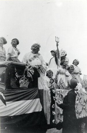 Photograph showing the side of the cab of a lorry and its back dressed as a float; on the side of the cab are Union Jacks, bunting and a crown; on the back of the lorry are a decorated cloth and five girls and two women dressed in light-coloured robes with flowers on them; a model of Britannia is also on the float; two girls are sitting on the top of the cab and a woman is standing on the ground watching; the occasion has been identified as the Coronation of George VI and the place Wingate