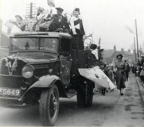 Photograph showing the bonnet and cab of a motor lorry with the registration F5649 driving towards the camera and decorated as a carnival float; man and a woman in Scottish dress can be seen standing behind the cab; the other figures on the lorry are indistinct; part of a procession of children can be seen behind the lorry and part of a crowd lining the street can also be seen; the photograph has been identified as Partridge Haulage Contractors Wagon in Coronation Day Celebration in Wingate